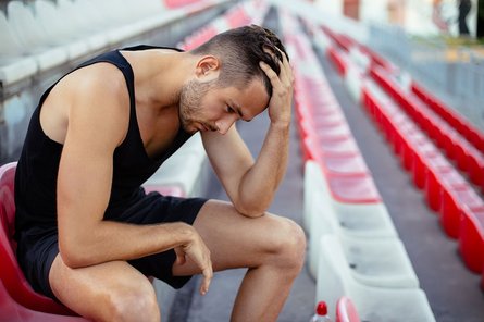 Ein Sportler sitzt auf einer Tribüne, hält die Hand an seinen Kopf und schaut erst auf den Boden