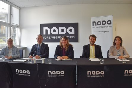 Fünf Personen sitzen an einem Konferenztisch der NADA vor einem NADA-Logo und einem Roll-up-Banner in einem hellen Besprechungsraum.