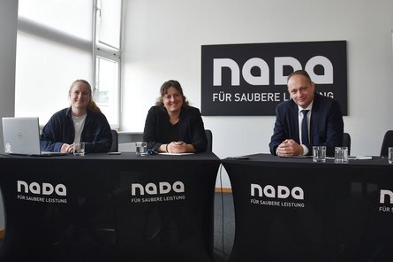 Drei Personen sitzen an Tischen mit NADA-Branding in einem Konferenzraum. Im Hintergrund ist ein NADA-Logo mit dem Schriftzug „Für saubere Leistung“ zu sehen.