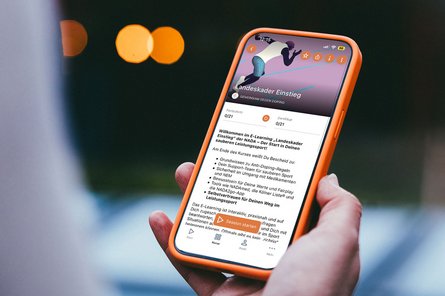 Hand hält Smartphone mit geöffneter E-Learning-App für Landeskader-Athletinnen und -Athleten mit Text und grafischer Darstellung einer laufenden Person